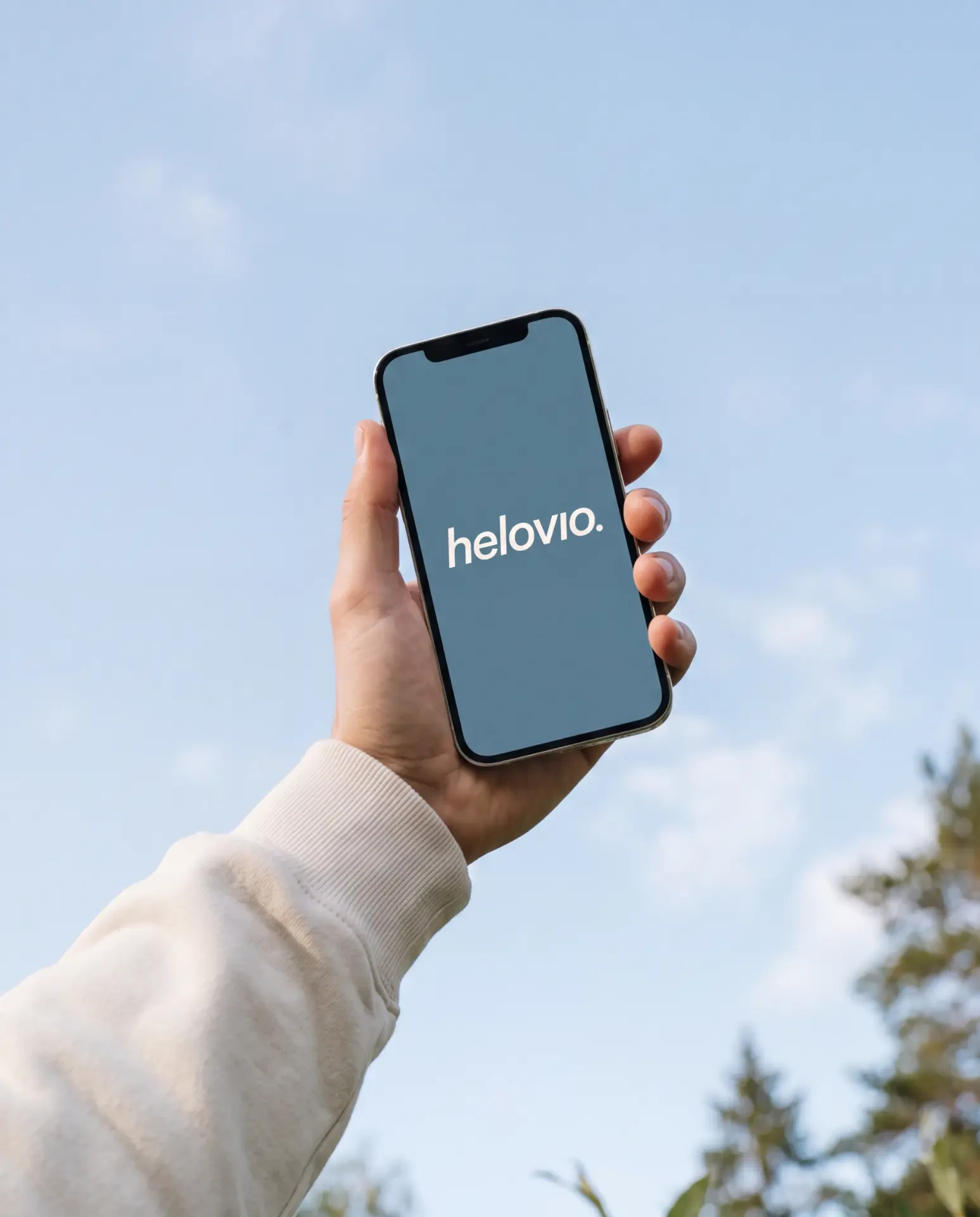 Hand håller en mobil med Helovios logotyp mot en klarblå himmel
